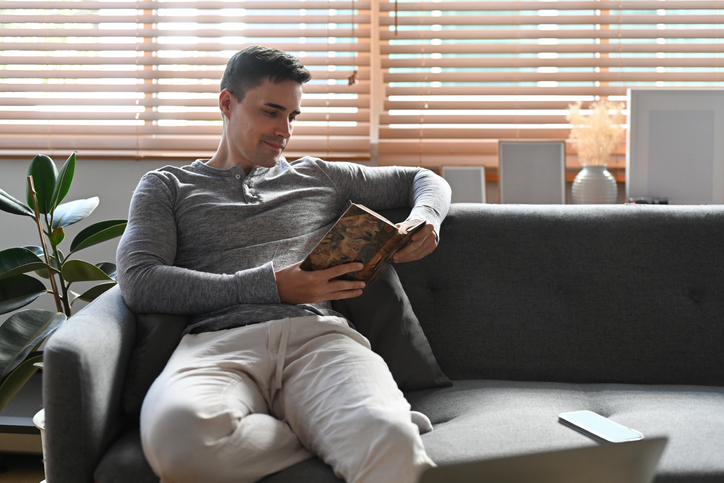 Handsome Caucasian man on a grey shirt sitting on a couch at home reading a book.