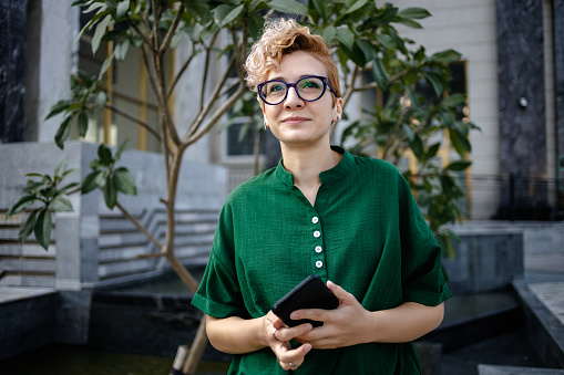 Lonely woman with asymmetric blonde hair wearing a green shirt with mandarin collar and glasses looking for someone while holding a smartphone in Hanoi, Vietnam