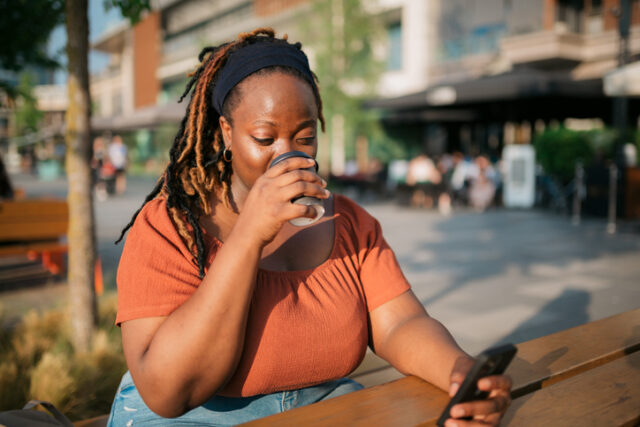 Young adult woman sitting in an outdoor coffee shop, using a smart phone and enjoying a cup of coffee