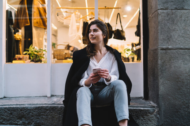 Caucasian female in long coat resting at shopping street with clothing wear on background using cellphone gadget on leisure, pondering woman with smartphone technology thoughtful looking away
