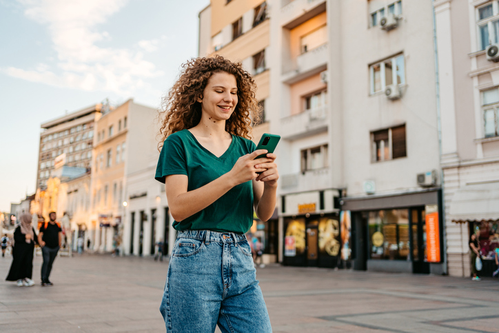 Beautiful young woman using smart phone while walking down the street on a sunny day.