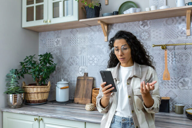 A sad young hispanic woman in the kitchen, pressing her phone and surprised by what she sees on her phone