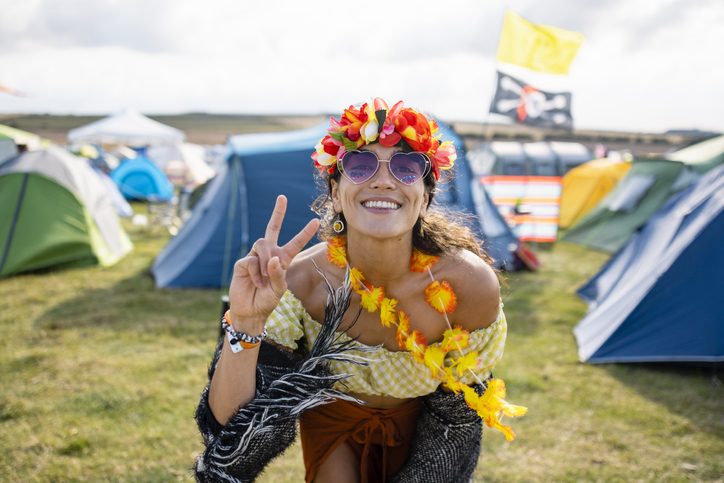 A young woman at a festival in Lindisfarne, North East England. She is wearing a flower headband, flower garland and heart sunglasses and doing the peace sign while looking and smiling at the camera.