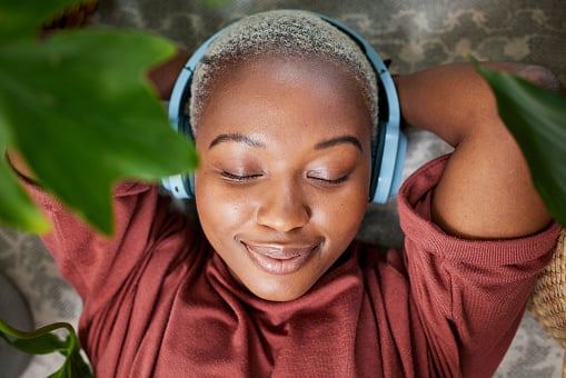Woman uses headphones to listen to music to relax on a floor with plants for peace and calm. Headshot of a black female person meditate with sound, audio and podcast or radio at home