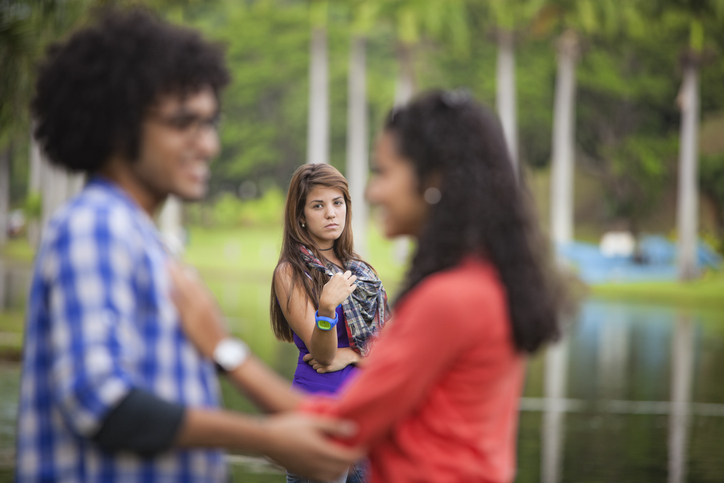 Young couple holding hands being observed by a woman in the background. Love triangle. Selective focus on woman in the background.