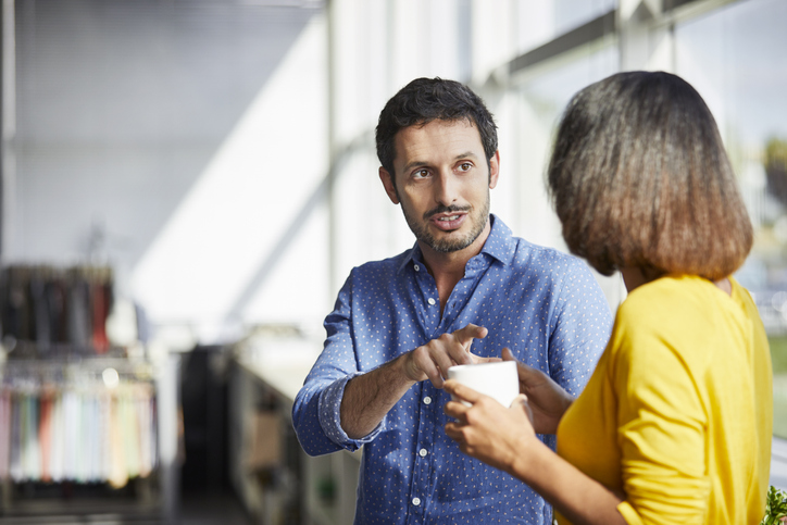 Confident businessman communicating with colleague having coffee. Male and female professionals are standing in office. They are discussing strategies at textile industry.