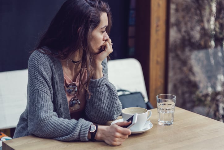 Brunette female sitting alone, holding her mobile phone, looking through the window and drinking coffee