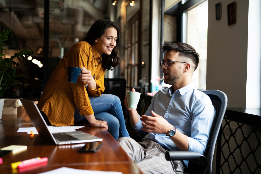 Two young colleagues in an office, the woman is sitting on the man's desk
