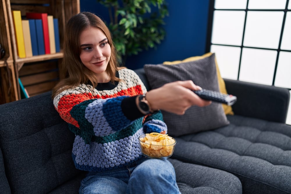 A young woman watching tv alone in the living room with a cup of popcorn and holding the remote