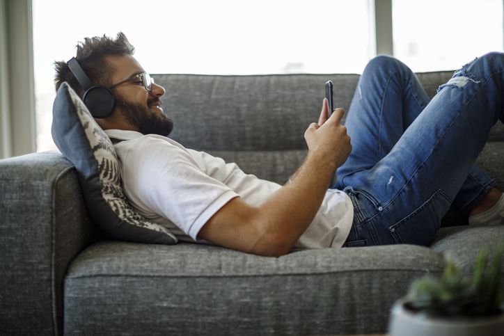 Smiling young man with wireless headphones using smart phone on sofa