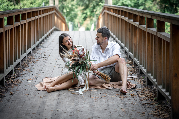 A beautiful girl and a young man sit on the bridge and enjoy communication, a date in nature, love story. The man is handing the woman a bunch of flowers