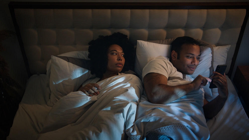 A woman laying next to her husband who is on his phone in bed.