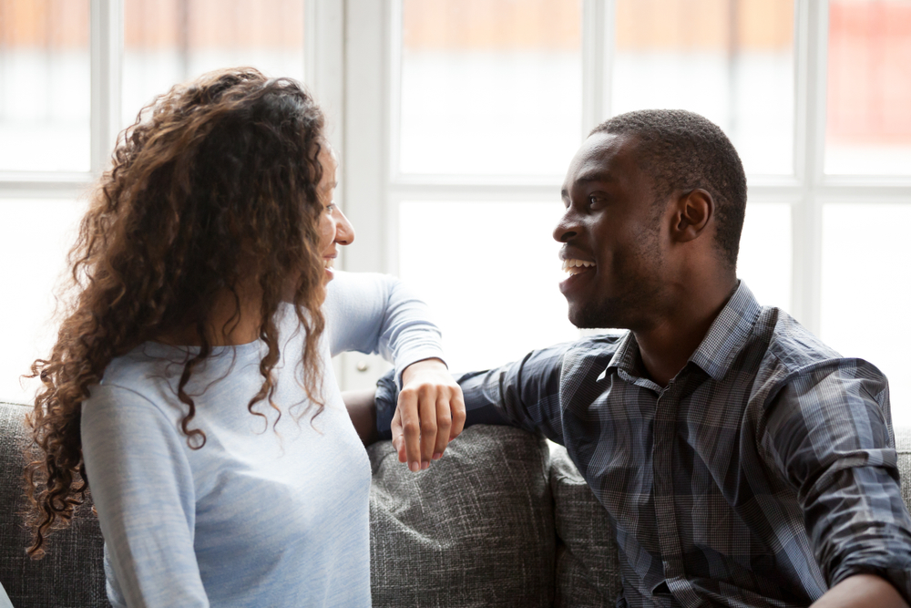 A black millennial couple sitting on the couch in the living room together, and talking to each other
