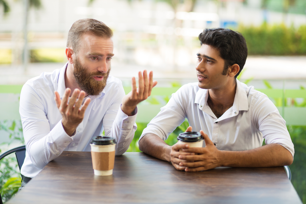 Two male friends talking and drinking coffee in an outdoor cafe.