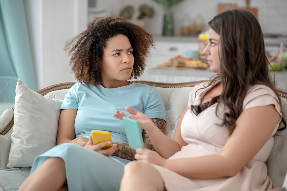 Two young ladies sitting on the couch and having a conversation, they are both holding their phones