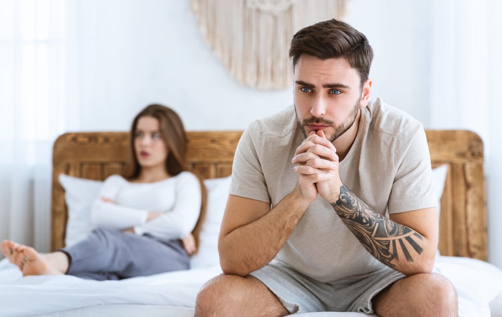 A sad man sitting in bed with his wife in the background
