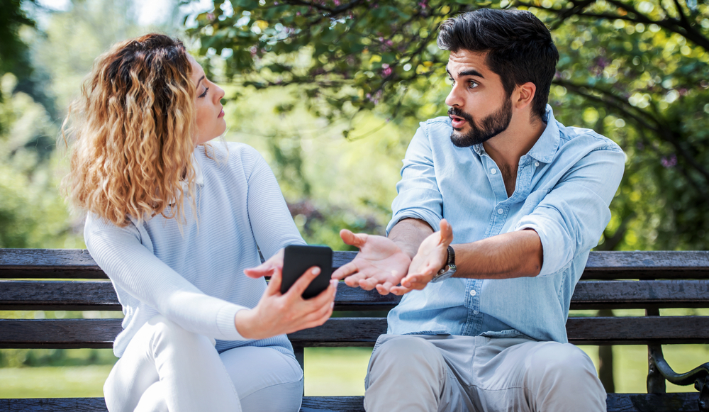 A young couple arguing and the man is pointing at the phone held by the woman