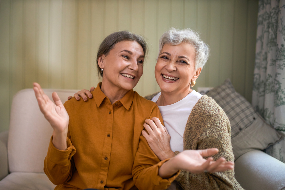 Two elderly women seated, holding each other and smiling