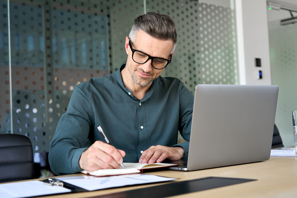 A busy middle age mature man writing in his notepad with his laptop in front of him