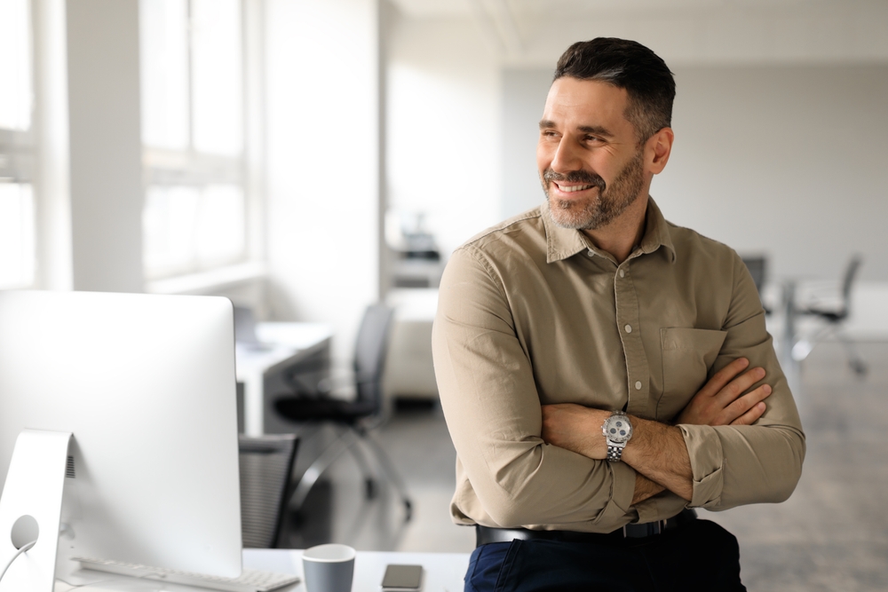 A happy middle age man at work and sitting on his desk with his hands folded