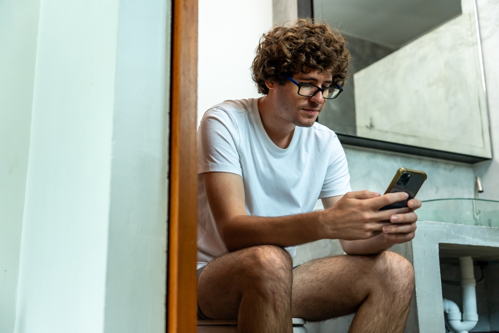 A young man sitting on toilet and using his phone