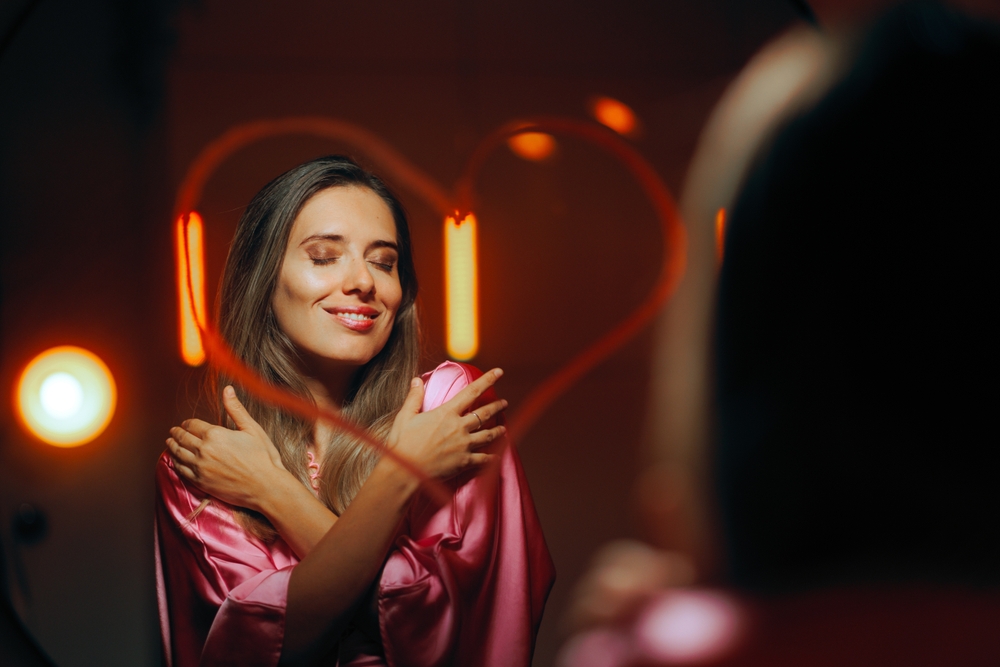 Woman looking in the mirror smiling and hugging herself in an act of self-love and confidence.