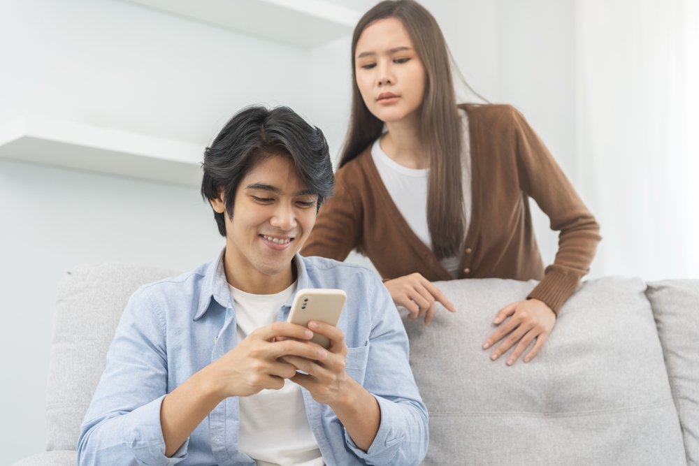 A suspicious woman trying to look into her husband's phone as he sits on the couch smiling and chatting