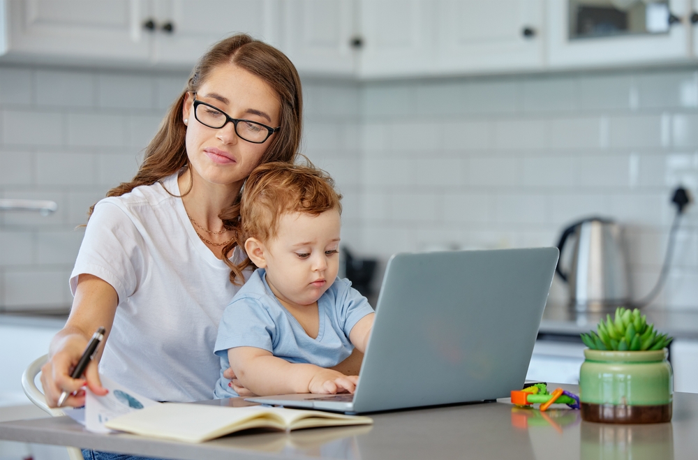 A mother sitting in front of her laptop, her young son is sitting on her lap and he is using the laptop, his mother is reading a book beside the laptop
