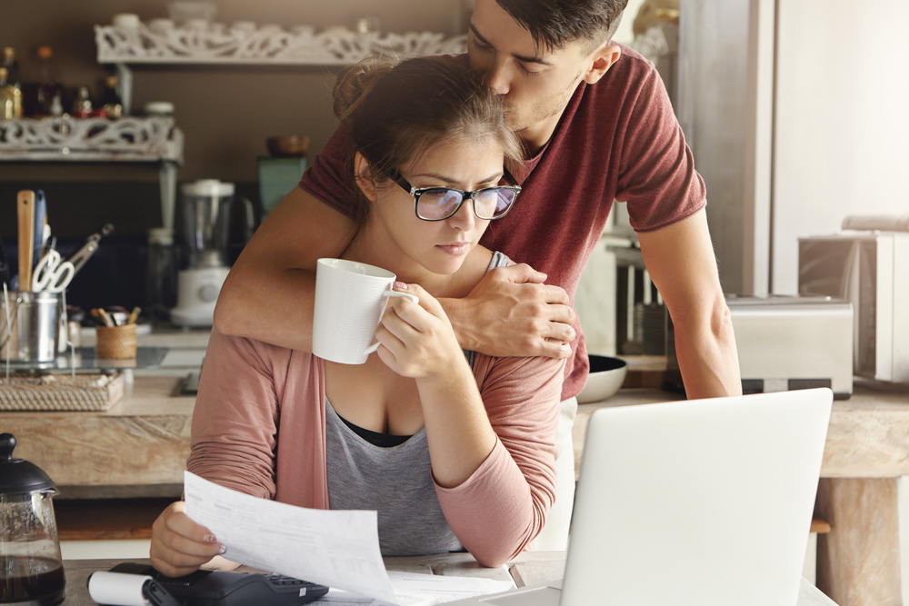 A young couple in the kitchen, the wife is going through the bills and the man comes from behind and kisses her head