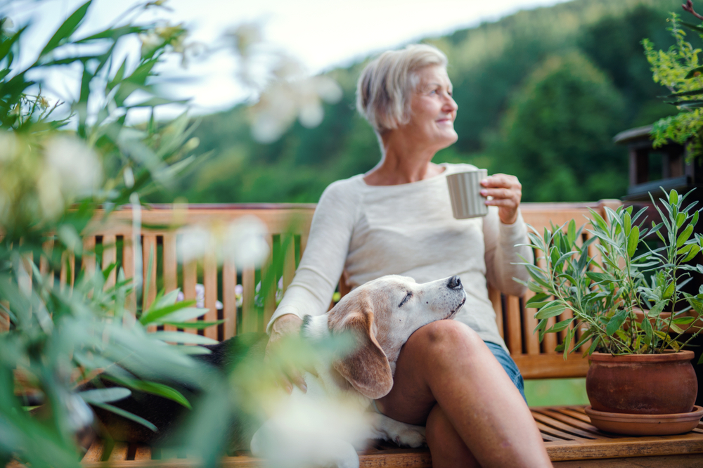 A senior woman sitting outside with a cup of coffee in her hands, and her dog resting his head on her knees