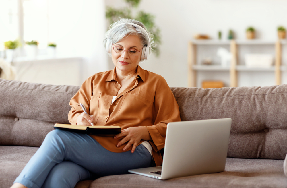 A mature woman using headphones and writing in her journal.