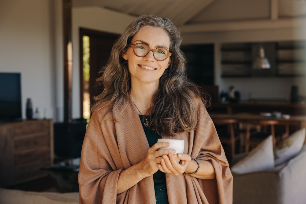 A happy senior woman standing in her house holding a cup of coffee and a blanket wrapped around her