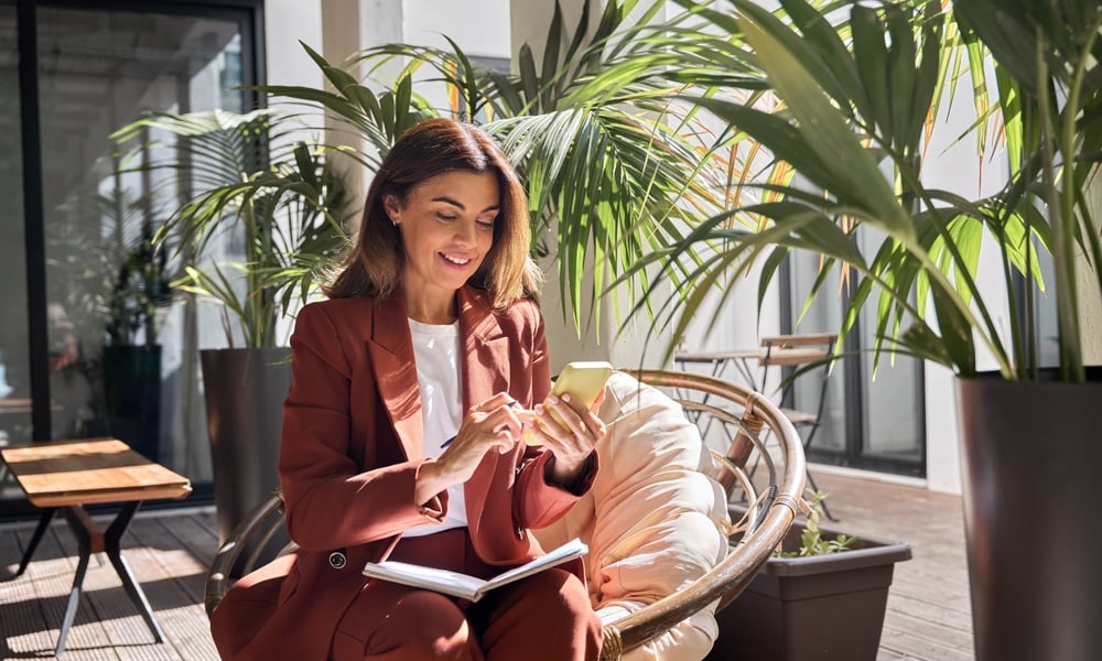 A smiling mature woman sitting by herself on a wicker chair outside while pressing her phone