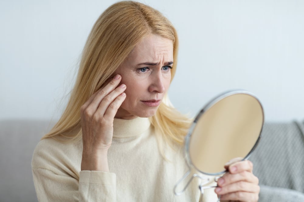 A middle aged woman looking at the mirror to check out her skin, she is unhappy with the signs of aging that she sees