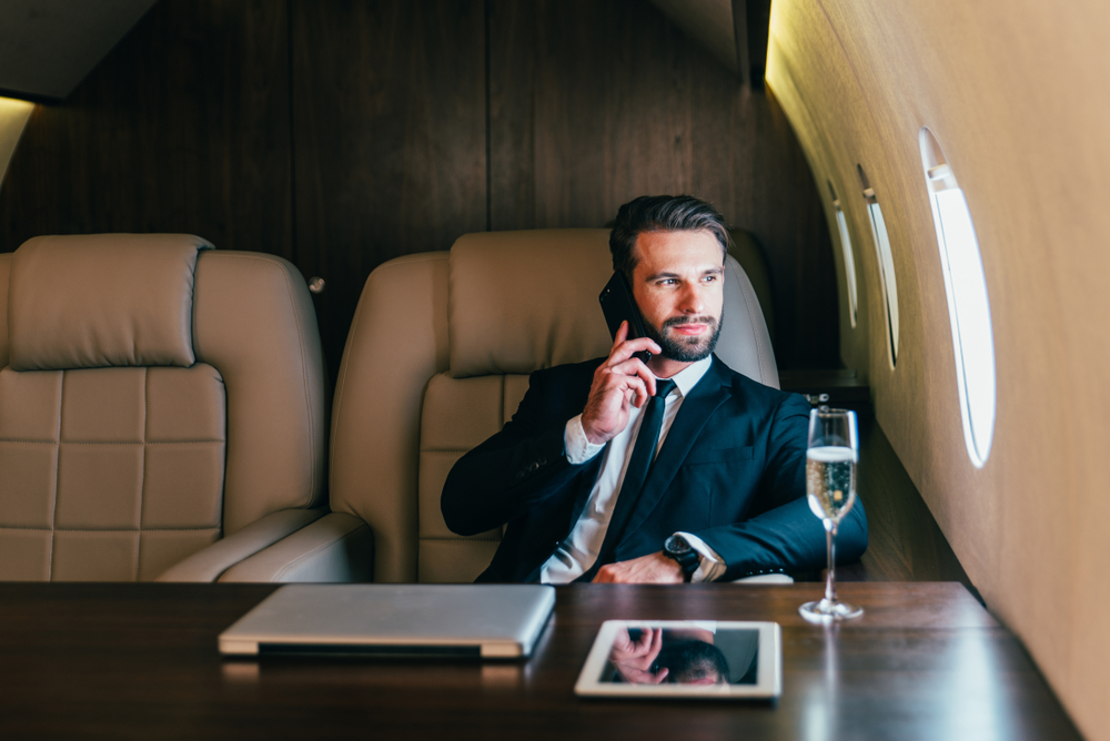 A businessman in a private jet while on his phone and enjoying a glass of wine