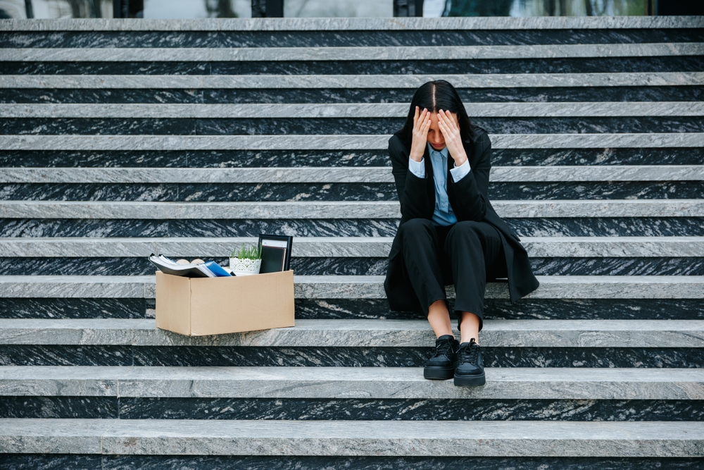 A woman sitting on the steps of her office building with a box containing items from her job desk after losing her job,