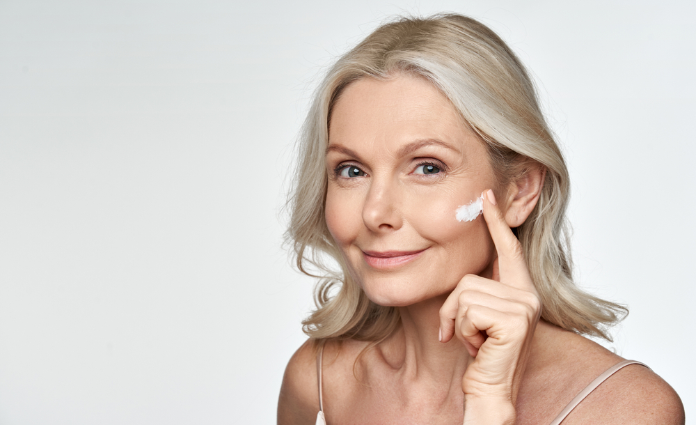 A middle aged woman applying skincare products on her face and smiling