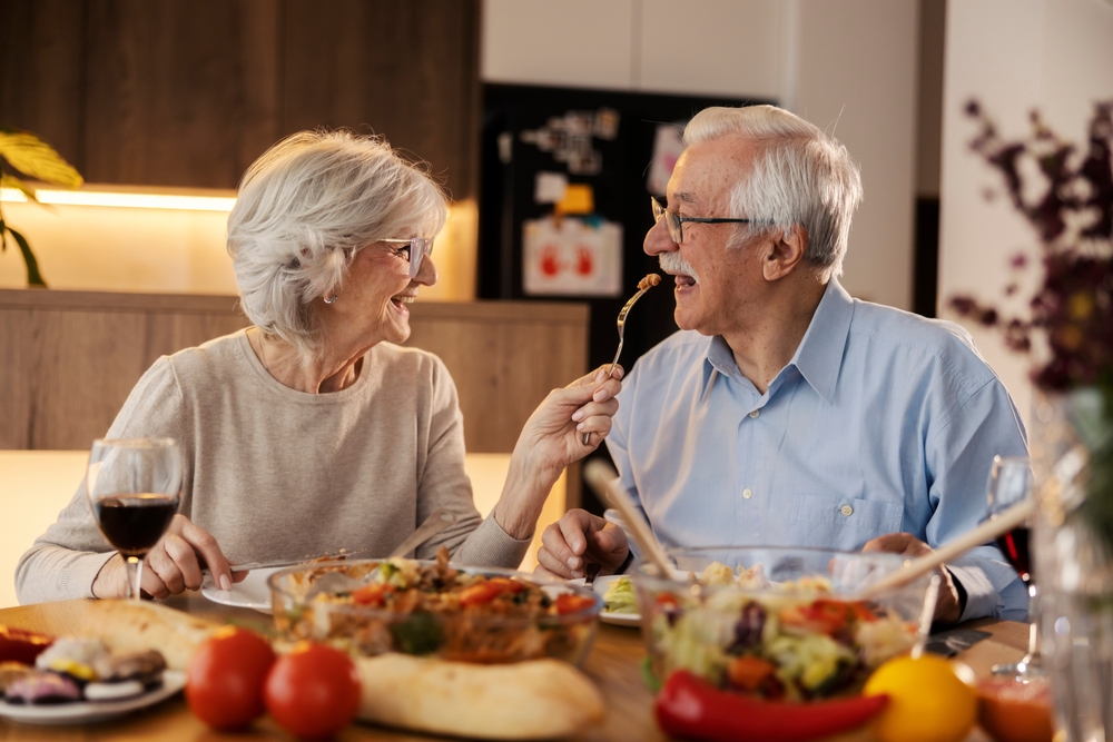 Healthy mature couple having lunch at the dinner table, the woman feeds the man with her fork