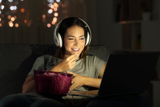 A woman watching a movie on her laptop alone, while using headphones and eating popcorn