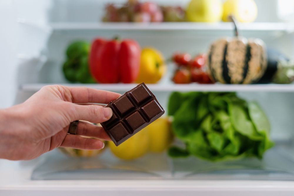 Fresh fruit and vegetables with a man holding dark chocolate near his refrigerator.