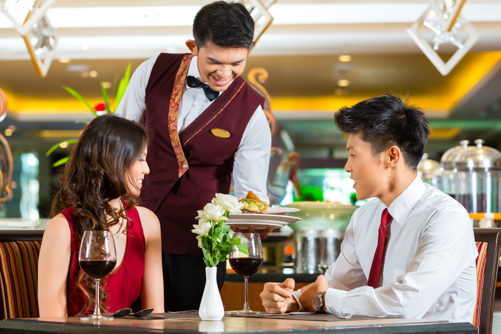 A young, happy lovely Asian couple out on a date in a fancy restaurant and they are being served by their young male waiter