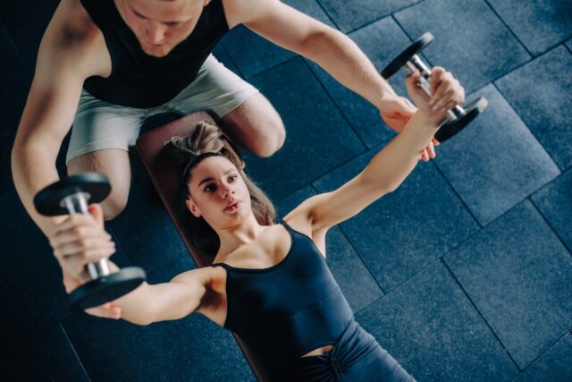 Male personal trainer guiding his female client, with weights in the gym.
