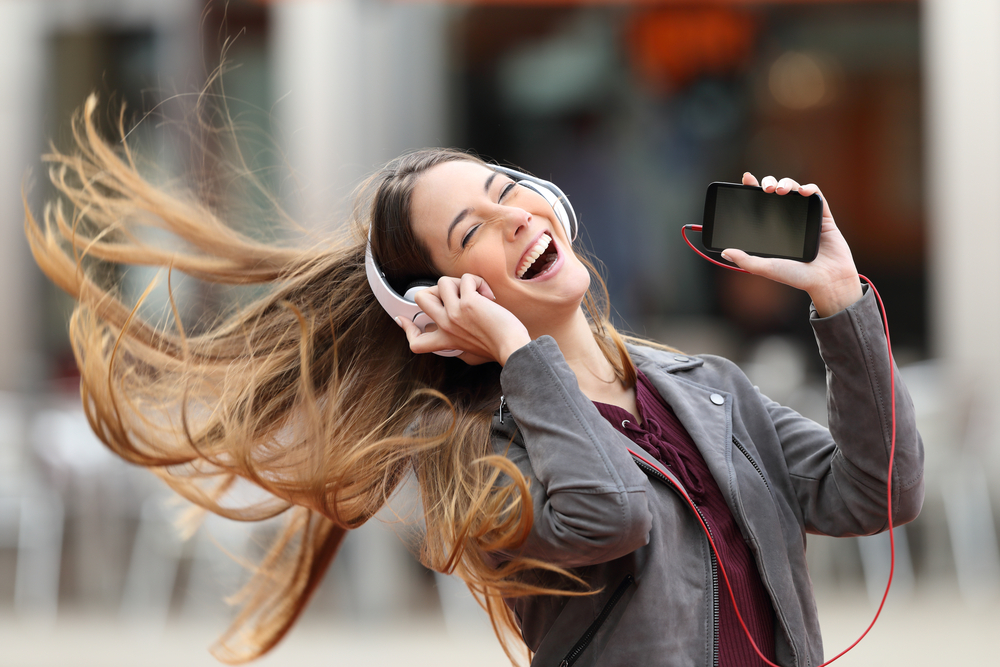 An excited laughing young woman dancing on the street while listening to music with her headphones and holding her smartphones