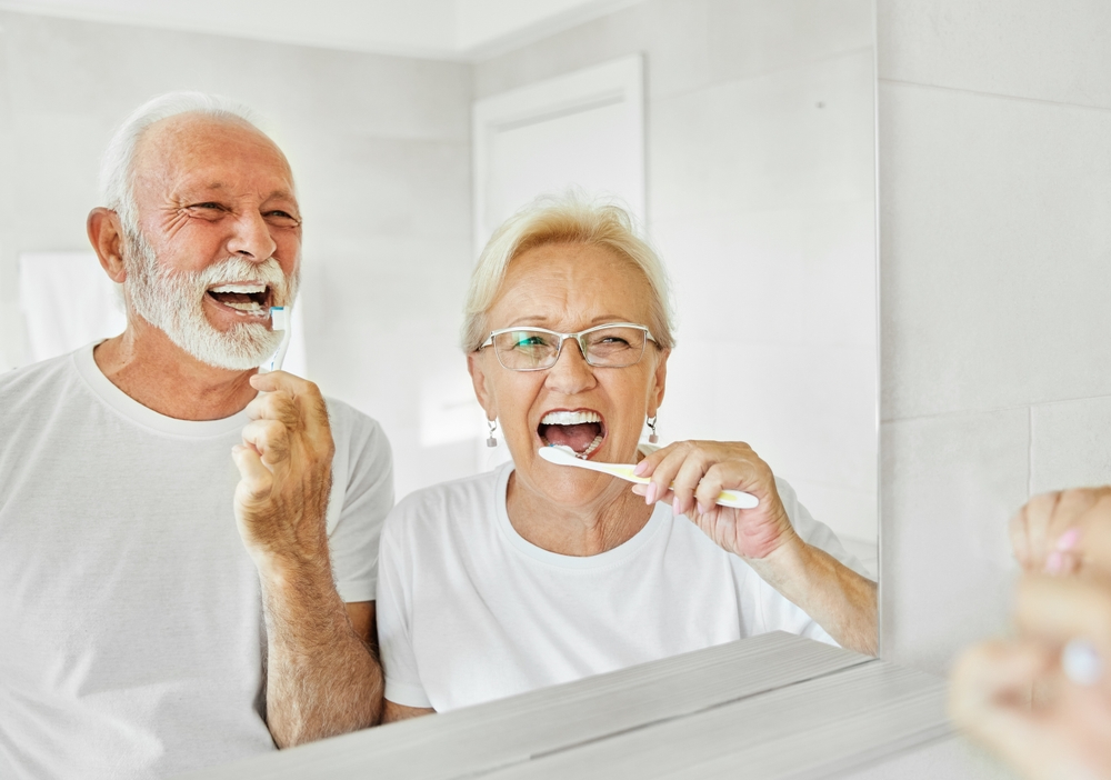 An elderly couple in the bathroom looking in the mirror as they brush their teeth.