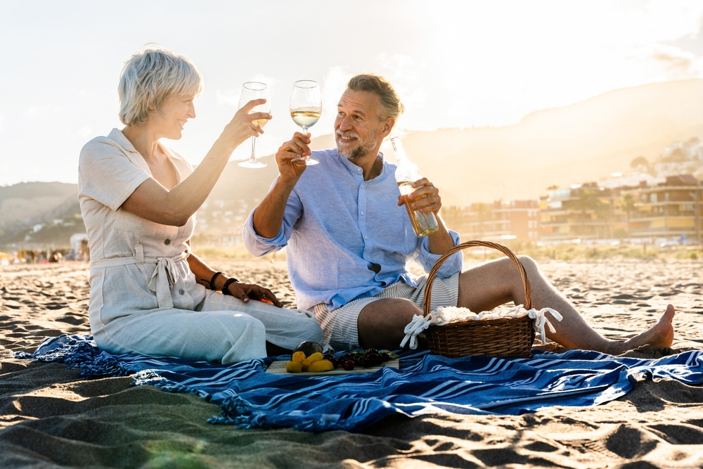 A happy mature middle age couple sitting on a blanket on the beach and enjoying a drink together