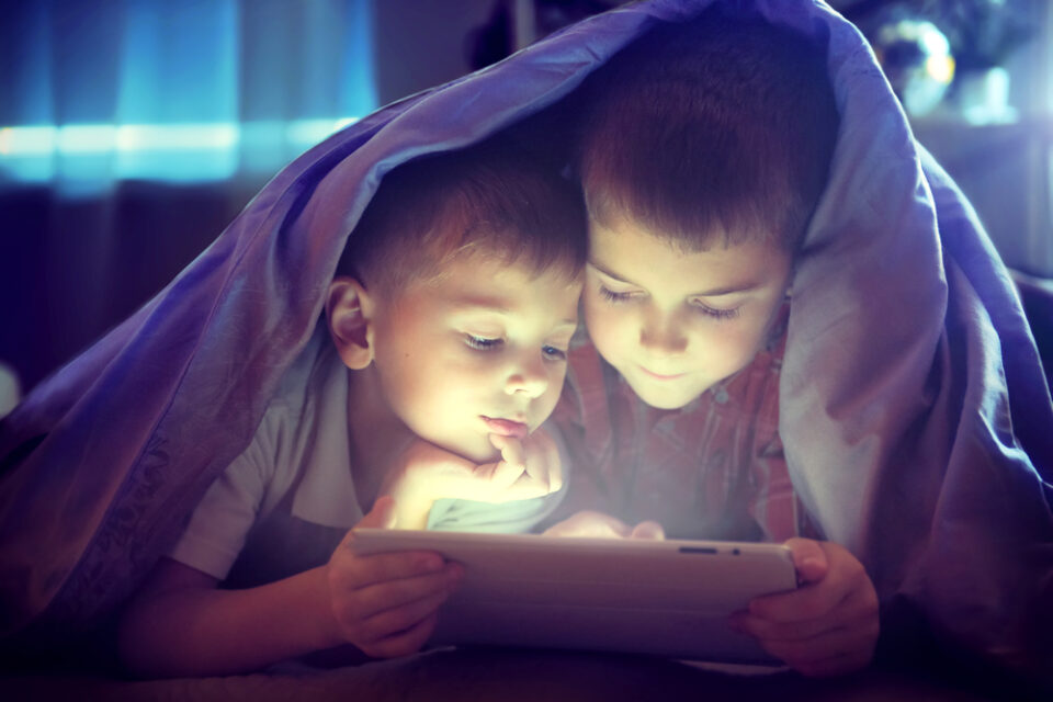 Two kids using their tablet under the blanket