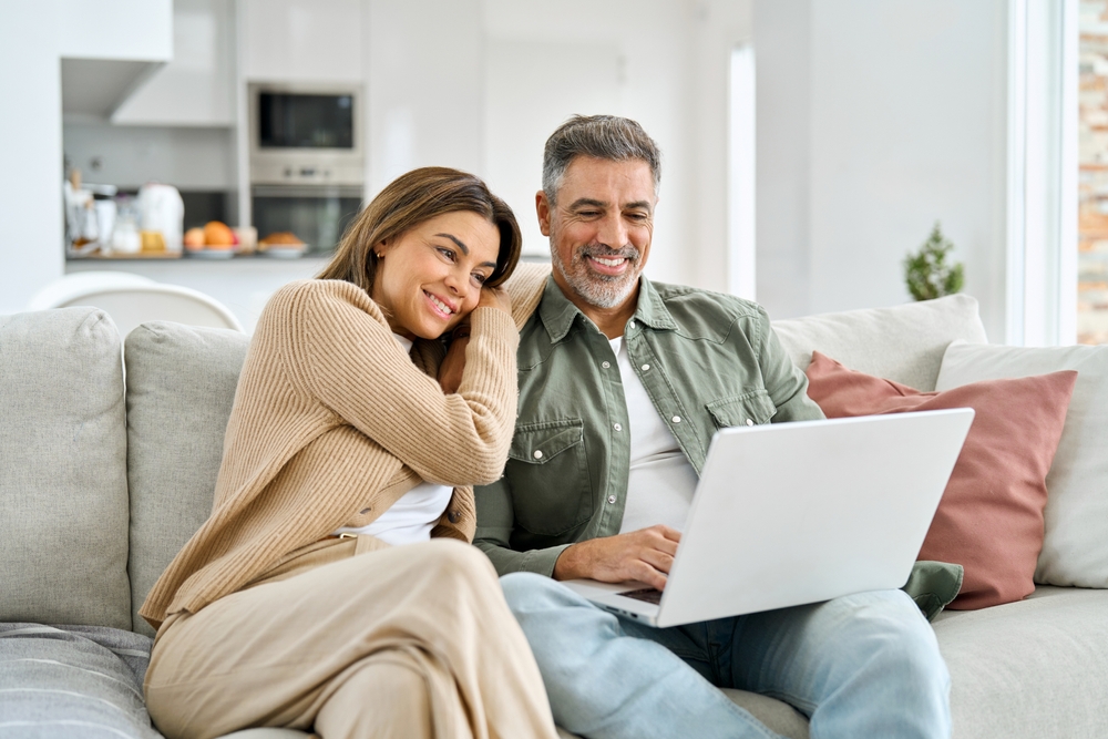 Happy middle aged couple sitting on the couch, smiling and looking at the laptop on the man's knee, the woman rests her head on the man's shoulder
