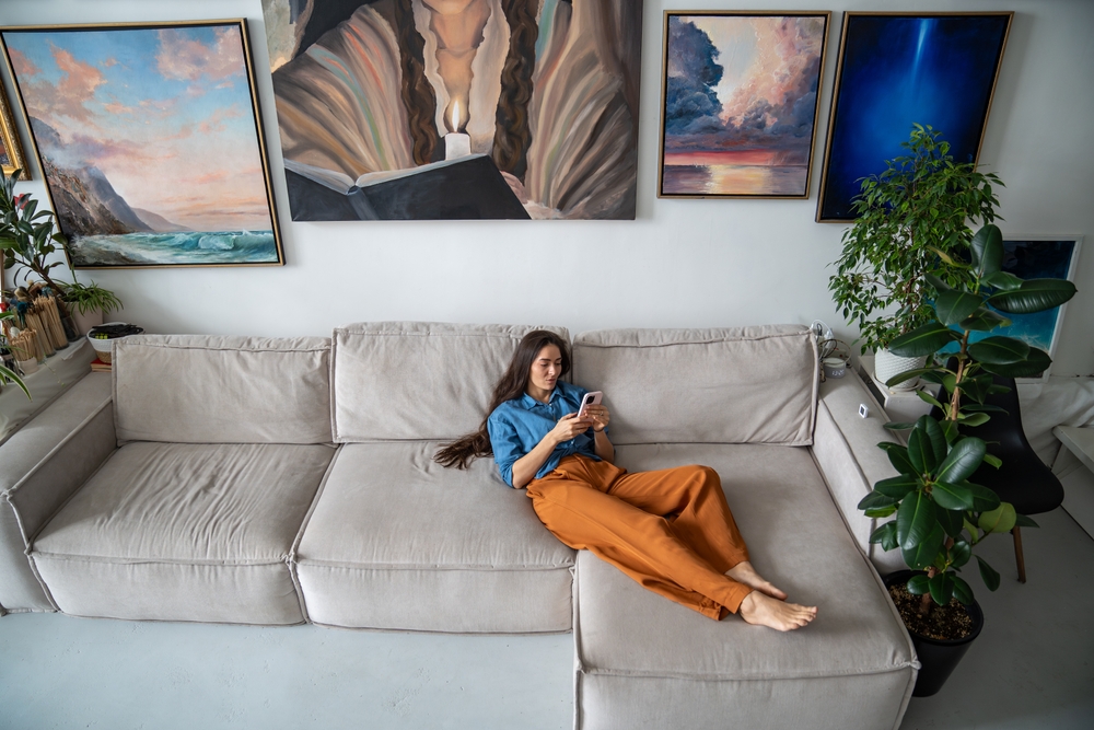 Lazy woman lying on her couch, pressing her phone, and procrastinating on doing chores